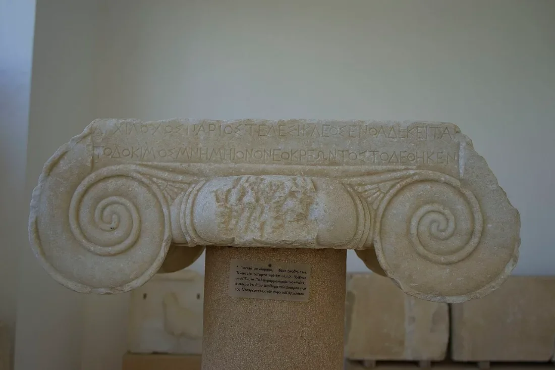 A marble Ionic capital with large volutes, on a pedestal in a museum.