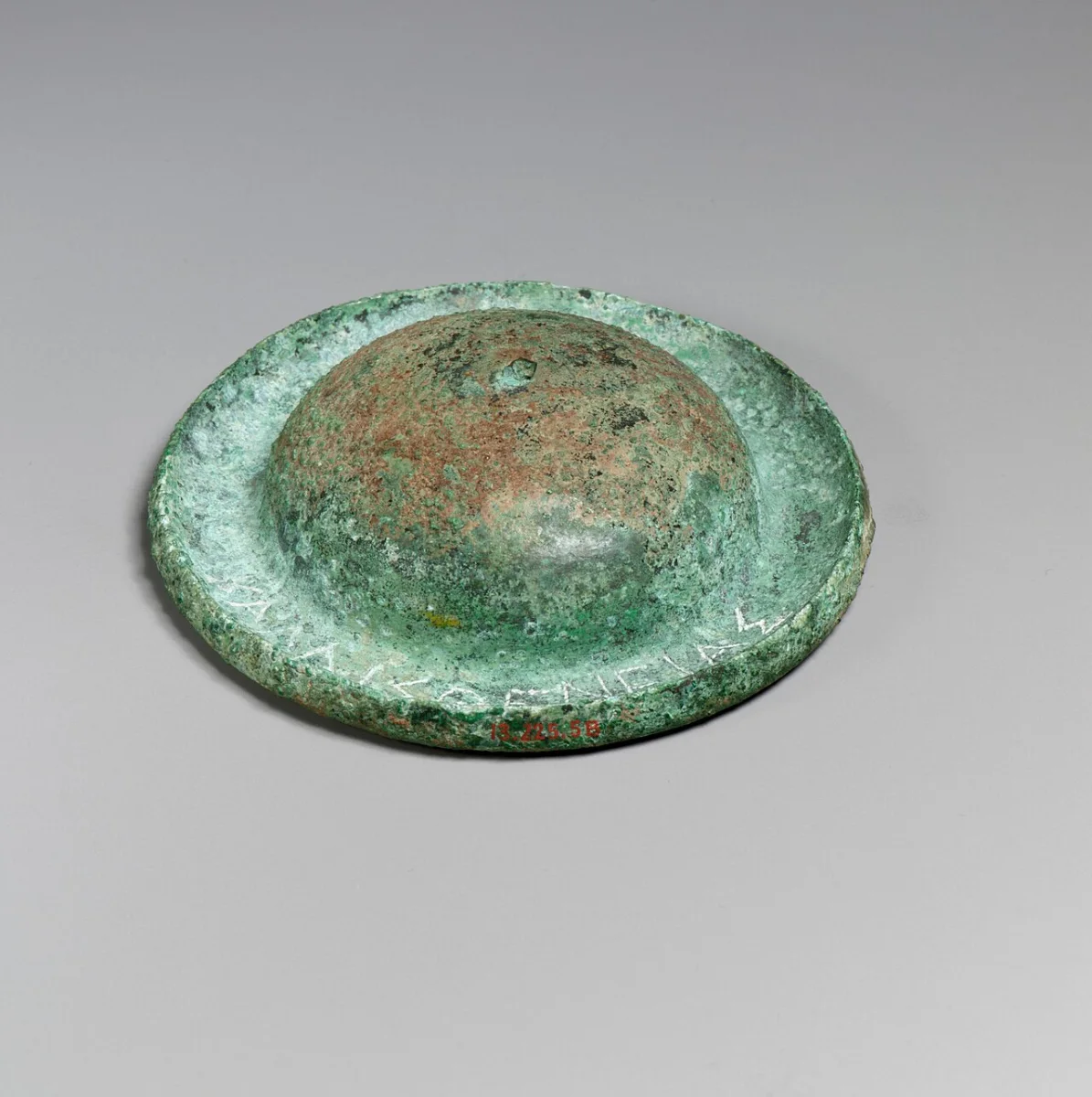 A small round bronze cymbal with a raised boss, green with age, seen from above.