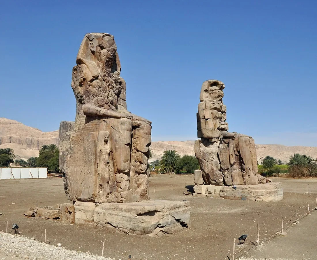 Two huge seated stone statues in a dusty plain.