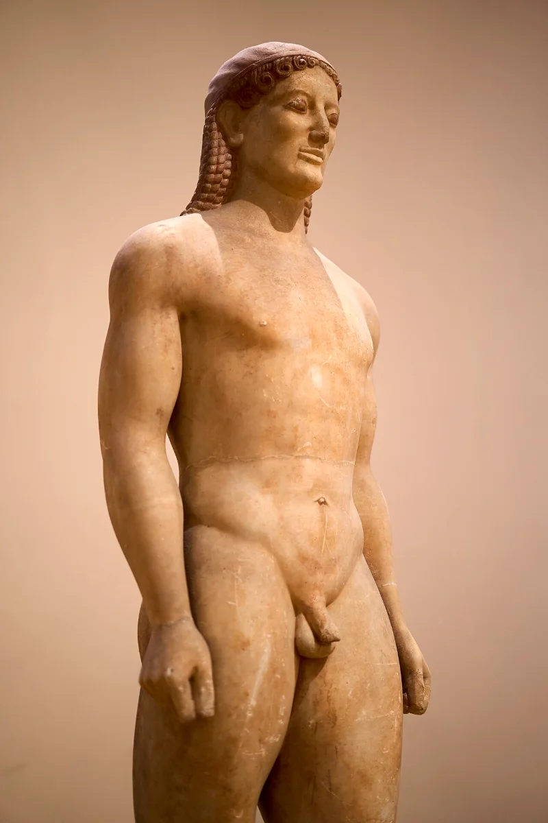 A marble statue of a naked young man standing with one foot forward, arms at his sides, long hair down his back.