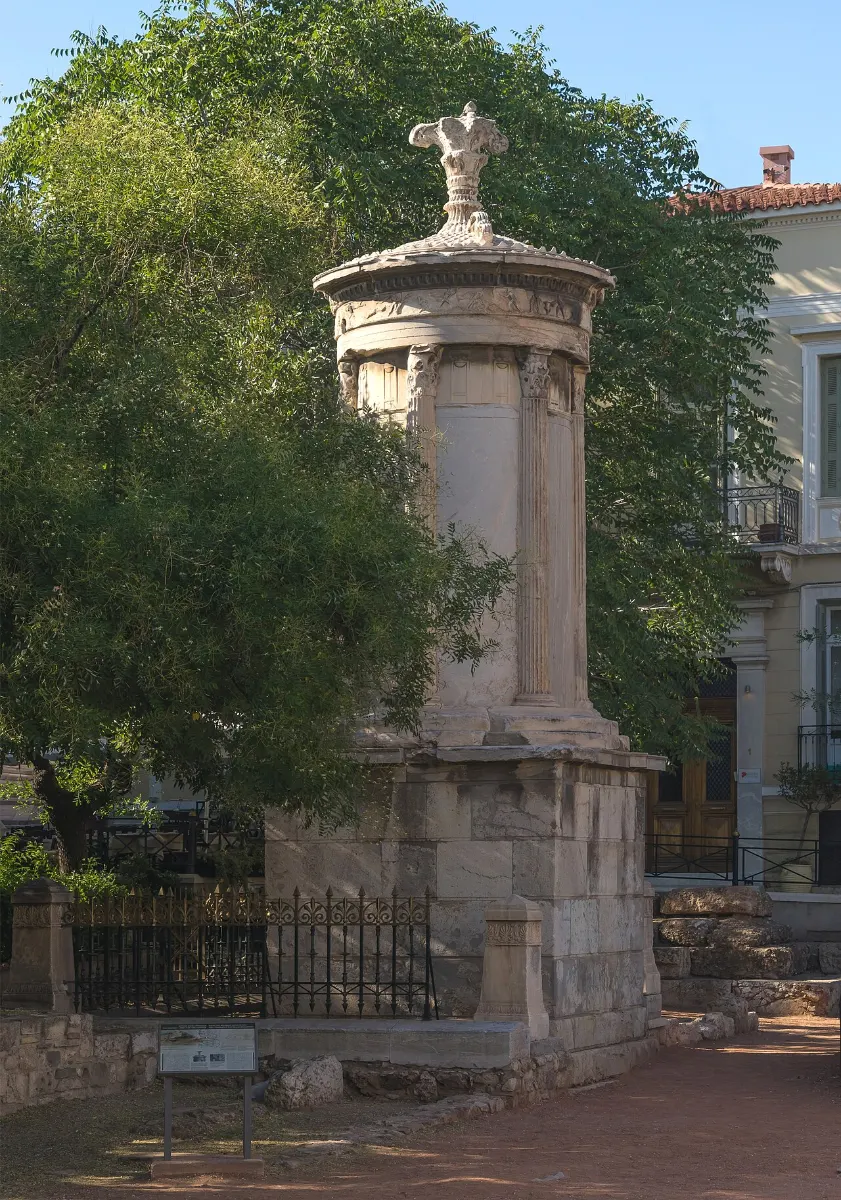 A slender round marble monument with Corinthian columns on a tall square base, crowned by a carved finial, among trees in Athens.