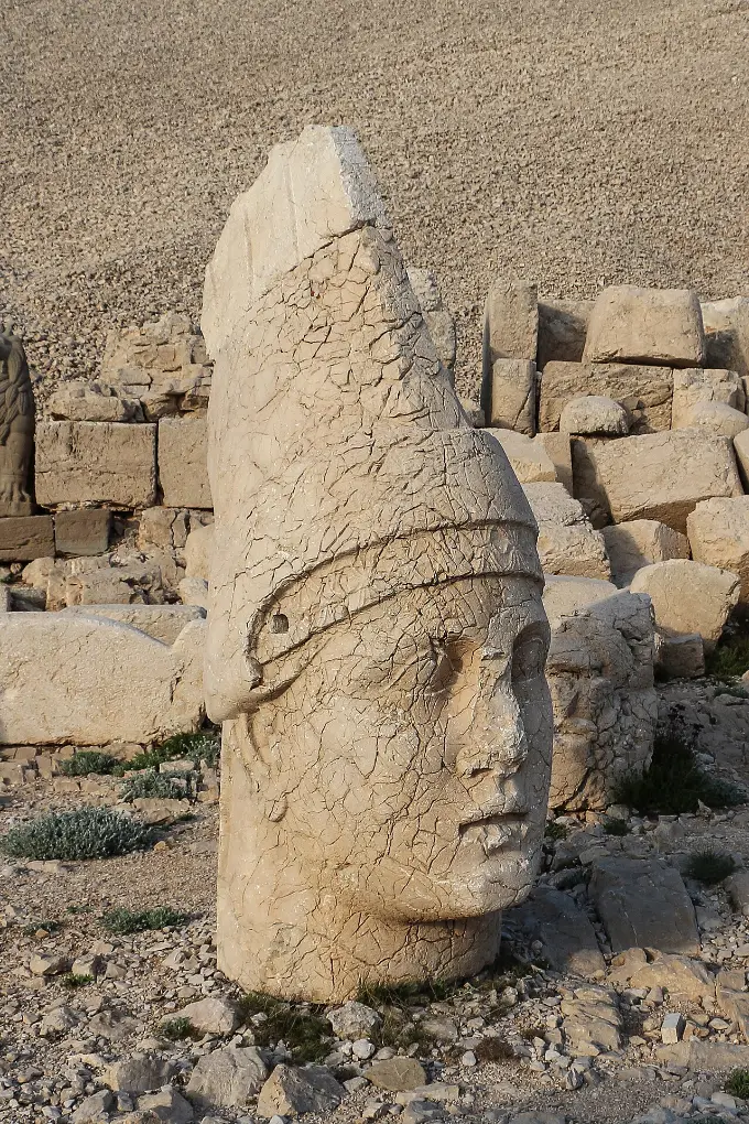 A colossal stone head in a tall peaked cap lying on a mountain terrace among fallen blocks.