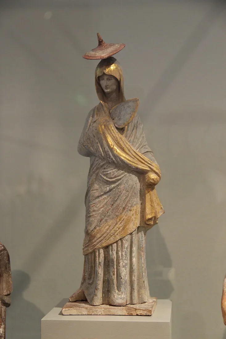 A painted clay statuette of a cloaked woman wearing a pointed sun hat.