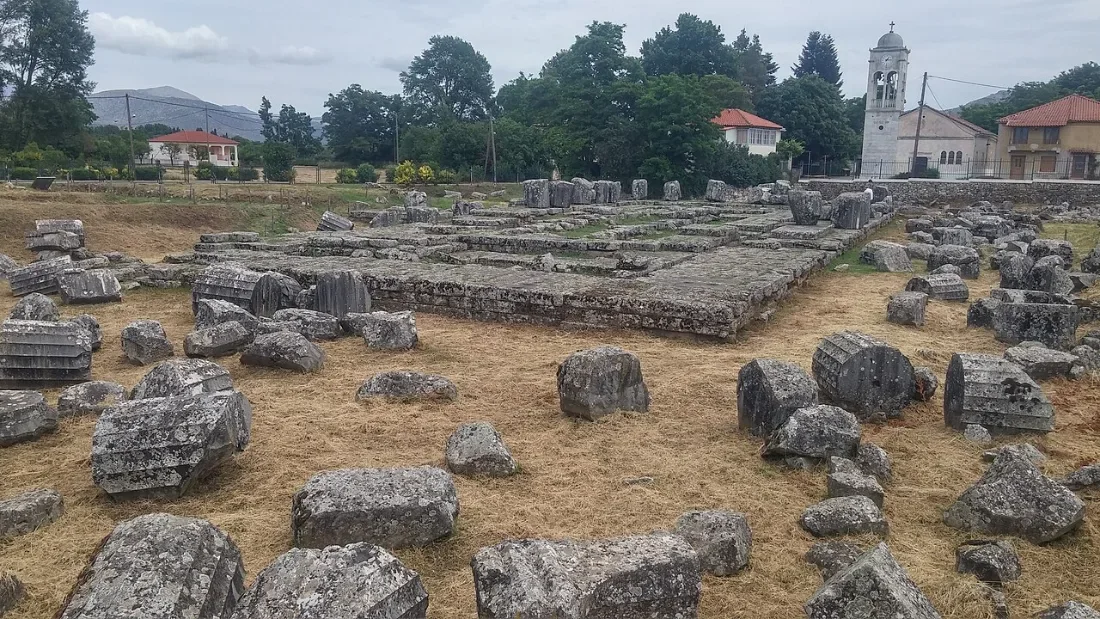 Scattered foundation blocks of a temple in a field, a village church behind.
