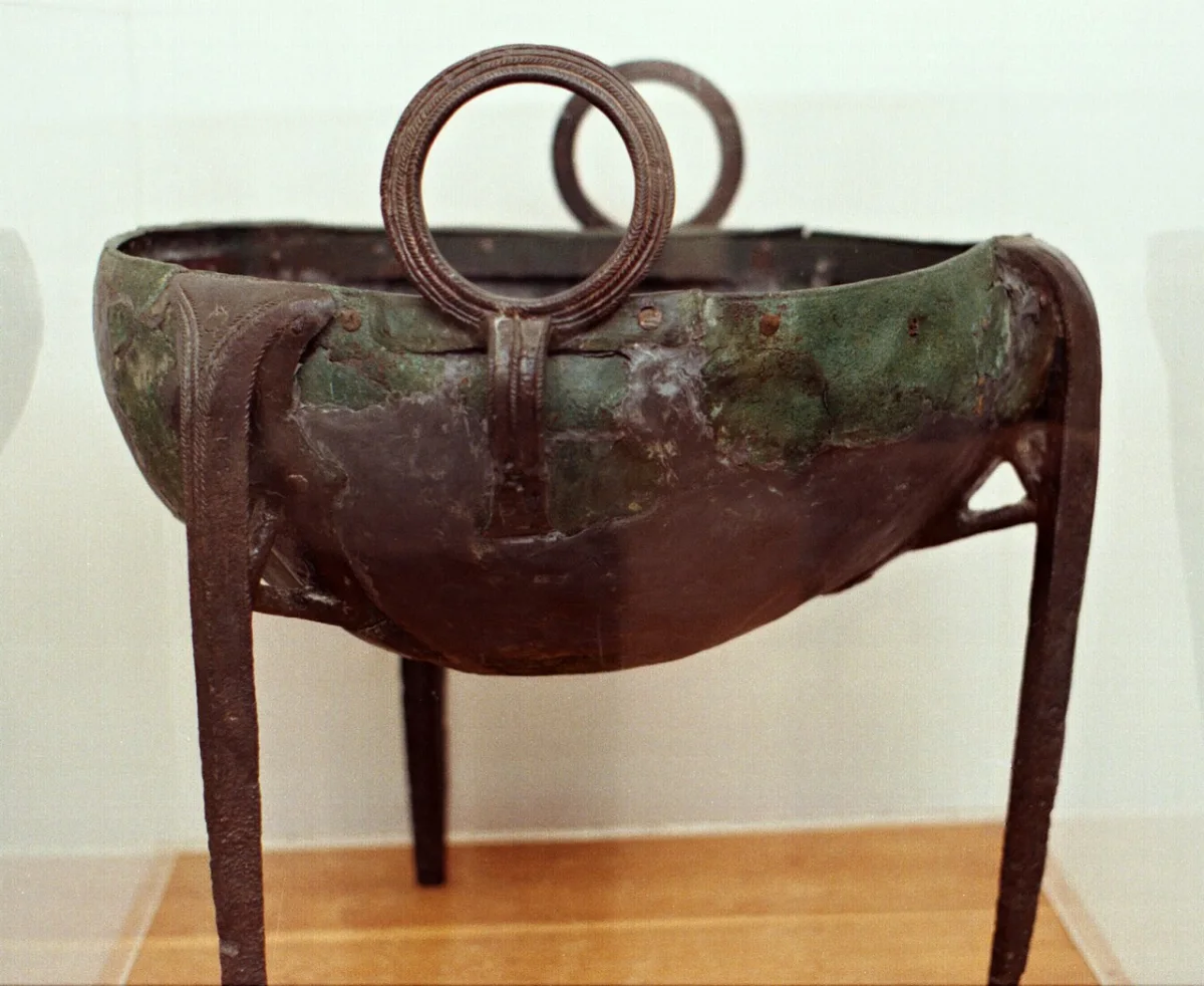 A large bronze cauldron on three legs, with two ring handles, in a museum case.