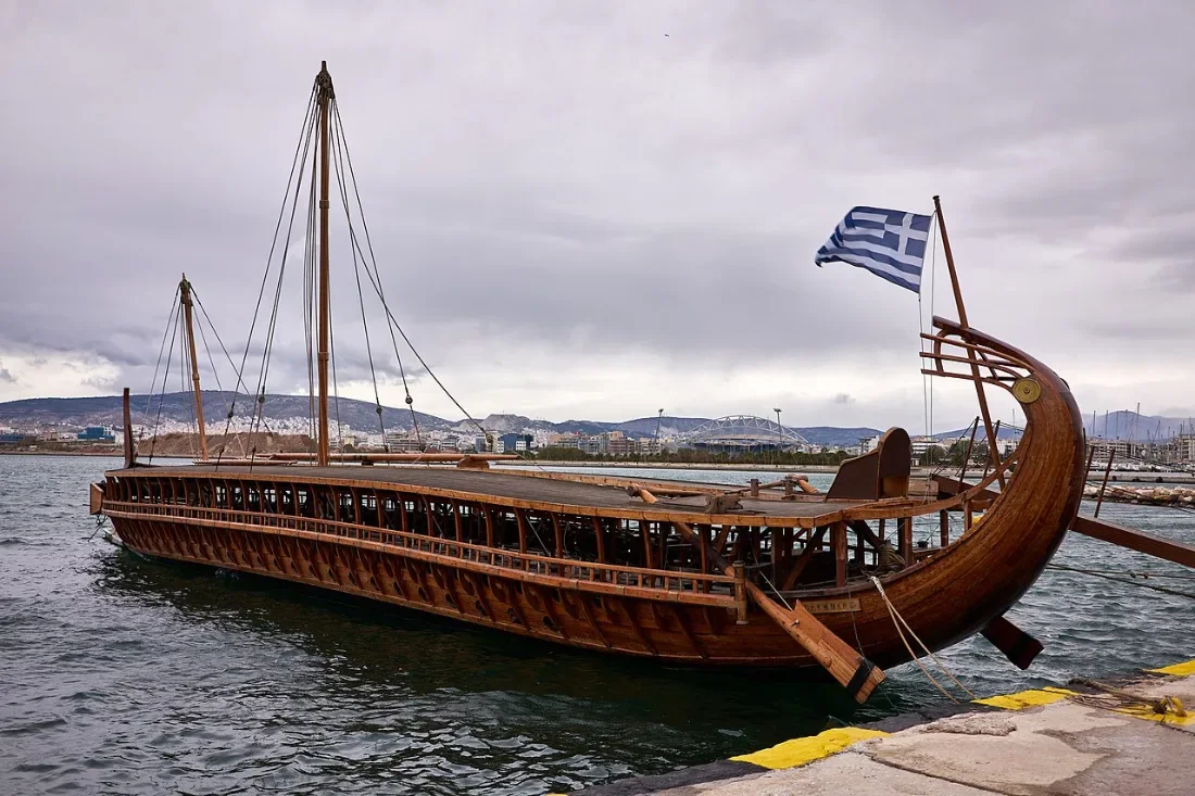 A long wooden warship with banks of oar ports and a raised stern, moored in a marina.